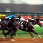 Jockeys in yellow, blue, and red silks urge their horses forward in a tightly bunched sprint down a dirt racetrack, with the lead horse just a nose ahead of the pack.