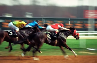 Jockeys in yellow, blue, and red silks urge their horses forward in a tightly bunched sprint down a dirt racetrack, with the lead horse just a nose ahead of the pack.