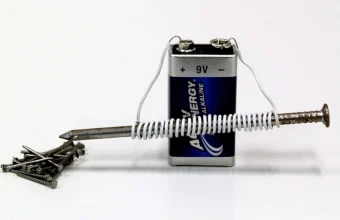 A homemade electromagnet made from a nail wrapped in wire and connected to a 9-volt battery, next to a small pile of nails it can pick up