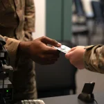 A U.S. servicemember hands a Common Access Card to another servicemember at an identity verification station with a biometric scanner