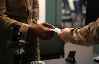 A U.S. servicemember hands a Common Access Card to another servicemember at an identity verification station with a biometric scanner