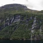 A steep green mountainside with a long waterfall above the dark water of Geirangerfjord in Norway