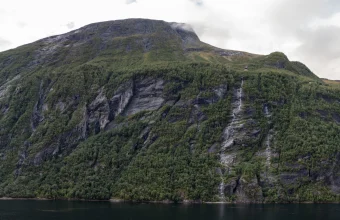 A steep green mountainside with a long waterfall above the dark water of Geirangerfjord in Norway