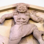 Close-up photograph of an archaic stone Gorgon relief sculpture from a Greek temple pediment, flanked by carved lion heads