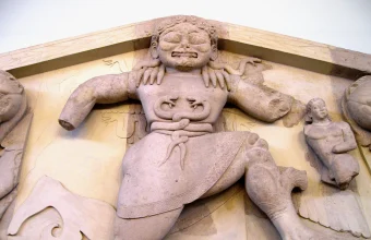 Close-up photograph of an archaic stone Gorgon relief sculpture from a Greek temple pediment, flanked by carved lion heads