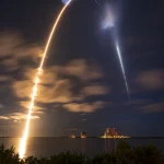 Long-exposure night photo of a SpaceX Falcon 9 rocket launch trail arcing over water at Kennedy Space Center