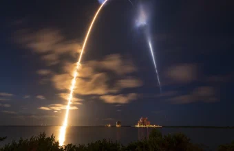 Long-exposure night photo of a SpaceX Falcon 9 rocket launch trail arcing over water at Kennedy Space Center