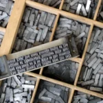 A composing stick holding set metal type spelling a pangram, resting on a wooden type case filled with metal sorts