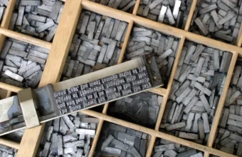 A composing stick holding set metal type spelling a pangram, resting on a wooden type case filled with metal sorts