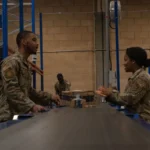 Two U.S. Air Force postal clerks sort packages on a warehouse conveyor belt, a physical stand-in for a work queue backlog