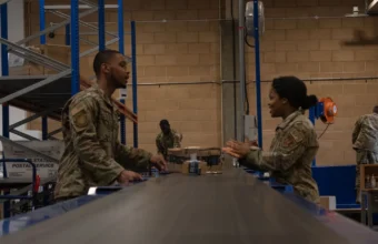Two U.S. Air Force postal clerks sort packages on a warehouse conveyor belt, a physical stand-in for a work queue backlog