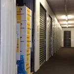 A storage unit hallway with one unit's roll-up door open and moving boxes stacked inside, while the row of units beyond it stay closed