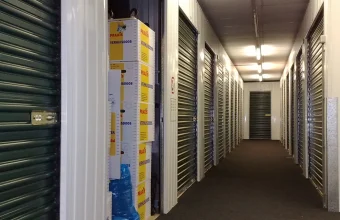 A storage unit hallway with one unit's roll-up door open and moving boxes stacked inside, while the row of units beyond it stay closed