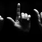 Three hands photographed in dramatic black-and-white lighting, spelling out letters in American Sign Language against a black background