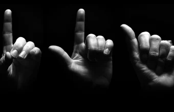 Three hands photographed in dramatic black-and-white lighting, spelling out letters in American Sign Language against a black background