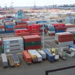Rows of stacked, multicolored shipping containers from different ocean carriers at a container terminal, with a city skyline in the background