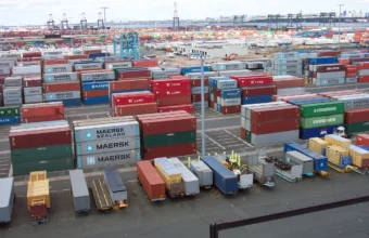 Rows of stacked, multicolored shipping containers from different ocean carriers at a container terminal, with a city skyline in the background