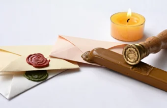A brass wax seal stamp and stick of sealing wax beside envelopes closed with red and green wax seal impressions, next to a lit candle