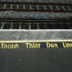 Yellow stenciled Irish text meaning 'Stay behind this line' painted beside a yellow boundary line at a railway platform edge, with tracks beyond