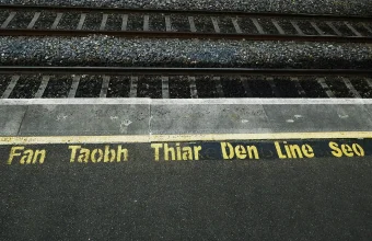 Yellow stenciled Irish text meaning 'Stay behind this line' painted beside a yellow boundary line at a railway platform edge, with tracks beyond