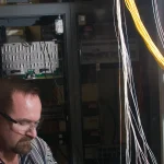 A technician handles bundles of orange and yellow fiber optic cables at a data center patch panel while splicing fiber on a workbench