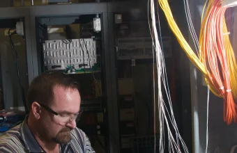 A technician handles bundles of orange and yellow fiber optic cables at a data center patch panel while splicing fiber on a workbench