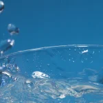 A clear glass tumbler of water overflowing and splashing at the rim against a blue background