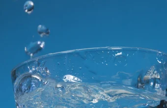 A clear glass tumbler of water overflowing and splashing at the rim against a blue background
