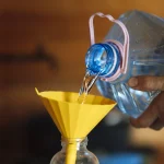 A hand pours water from a large jug through a wide yellow funnel into a narrow glass bottle