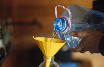 A hand pours water from a large jug through a wide yellow funnel into a narrow glass bottle