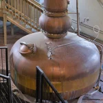 A gleaming copper pot still with a bulbous body and tall neck inside a distillery, used as a visual metaphor for AI model distillation