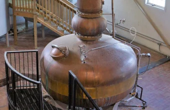 A gleaming copper pot still with a bulbous body and tall neck inside a distillery, used as a visual metaphor for AI model distillation