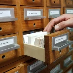 A hand reaching into an open library card catalog drawer to pull out a specific index card
