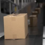 A single cardboard package on a warehouse conveyor belt with more packages queued behind it
