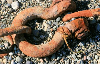 A massive rusted anchor chain shackle lying on a pebble beach, its iron surface deeply corroded orange and brown