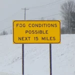 A yellow highway sign reading Fog Conditions Possible Next 15 Miles beside a snowy road