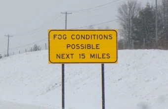 A yellow highway sign reading Fog Conditions Possible Next 15 Miles beside a snowy road