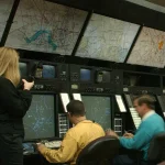 Air traffic controllers at consoles in a control room, each monitoring a separate screen while a supervisor stands overseeing the room