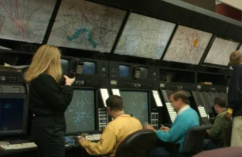 Air traffic controllers at consoles in a control room, each monitoring a separate screen while a supervisor stands overseeing the room