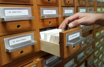 A hand reaching into an open library card catalog drawer to pull out a specific index card