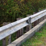 Galvanized steel guardrail bolted to wooden posts along the edge of a bridge approach, with a grassy verge and a gravel road beside it