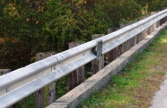 Galvanized steel guardrail bolted to wooden posts along the edge of a bridge approach, with a grassy verge and a gravel road beside it