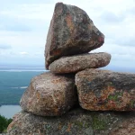 A stone cairn trail marker stacked on a mountain summit overlooking a lake and forest