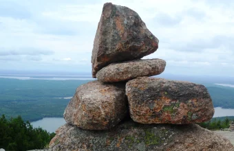 A stone cairn trail marker stacked on a mountain summit overlooking a lake and forest