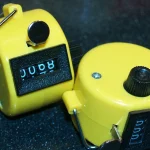 Two yellow mechanical tally counters resting on a dark speckled surface, one showing a four-digit count