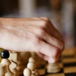 A hand reaches among a cluster of white chess pawns to move one piece on a wooden board, evoking a scheduler selecting which lower-priority workload to evict.