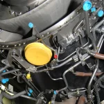 A dense wiring harness of tubes, hoses, and color-coded connector caps wrapped around a jet engine, with a technician working on the connections below