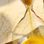 Golden sand flowing through the neck of a brass hourglass, pooling in the bottom bulb
