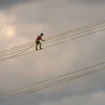A lineman walks along the conductor bundle of a high-voltage transmission line, performing maintenance directly on the live line carrying the load