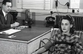 1945 black-and-white photograph of a polygraph lie detector test, with an examiner writing notes at a desk while a seated woman is wired to the machine